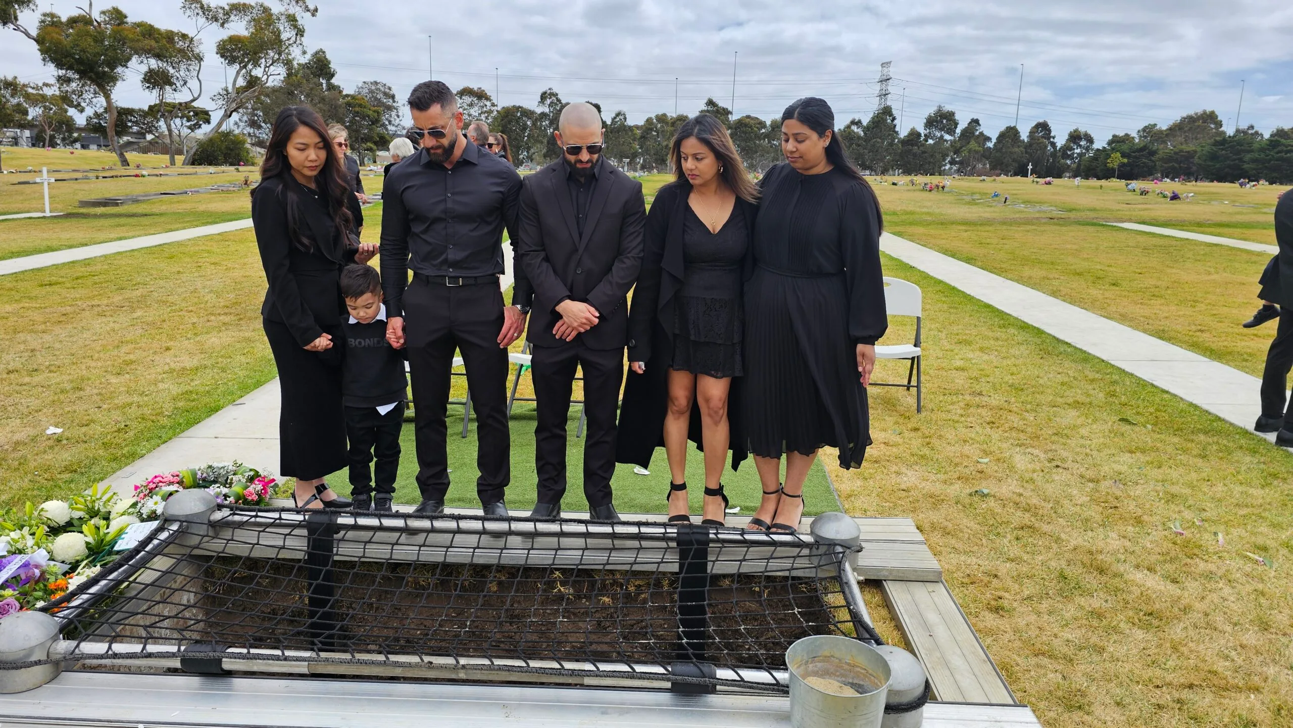Three women dressed in black and two men in black suits standing over a grave with flowers in the corner.