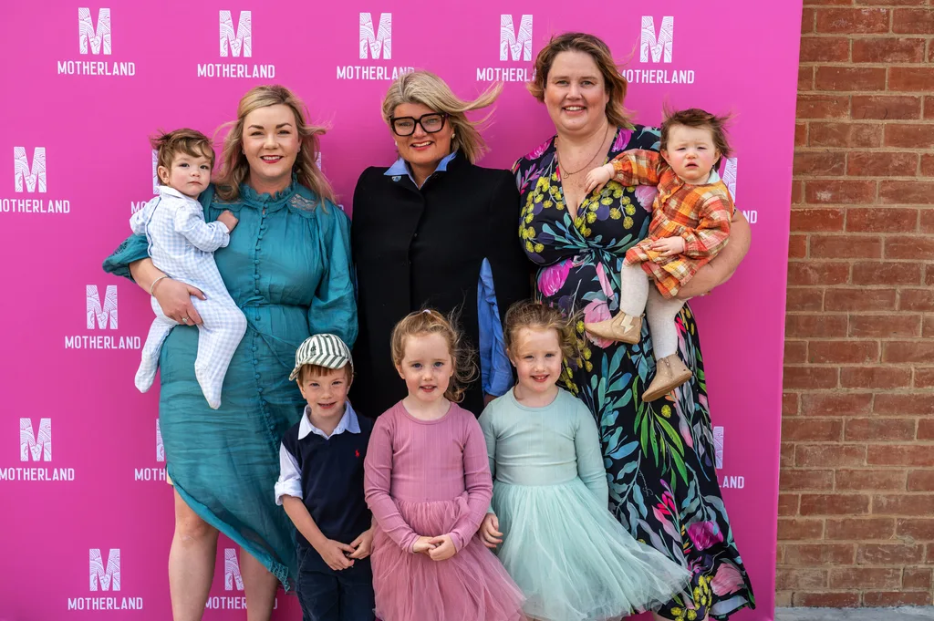 three women in black m, green and pattern dresses, two women holding babies and three chidlren standing one boy in hat and girl in pinik dress and girl green dress in front pink backgournd