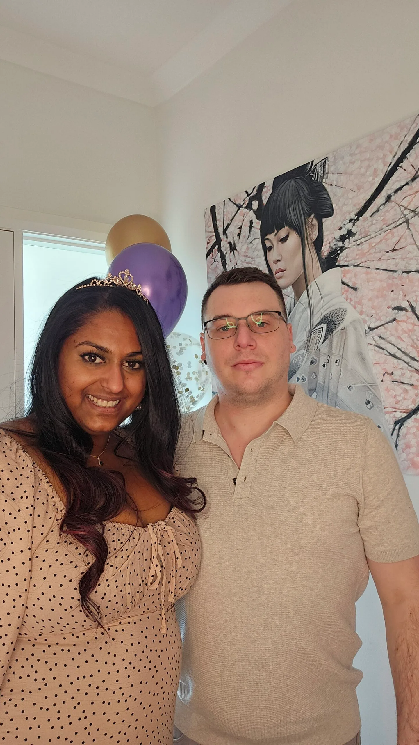 Left: Woman with black hair wearing brown dress and a tiara, smiling. 
Right: Man with short brown hair, wearing glasses and a grey shirt, wrapping arm around woman. Both smilling.
