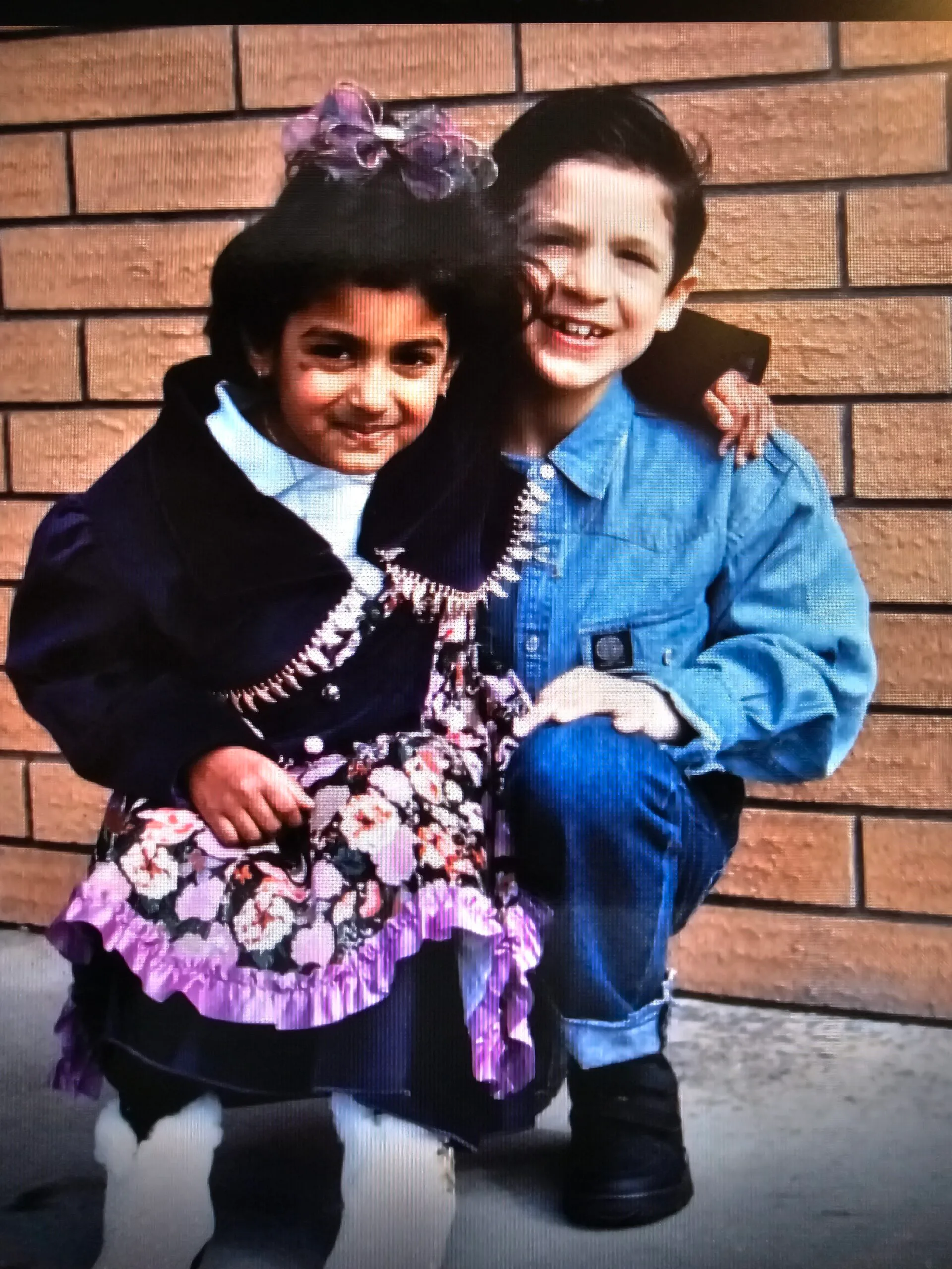 Little girl with black hair wearing purple dress wrapping arm around little boy.