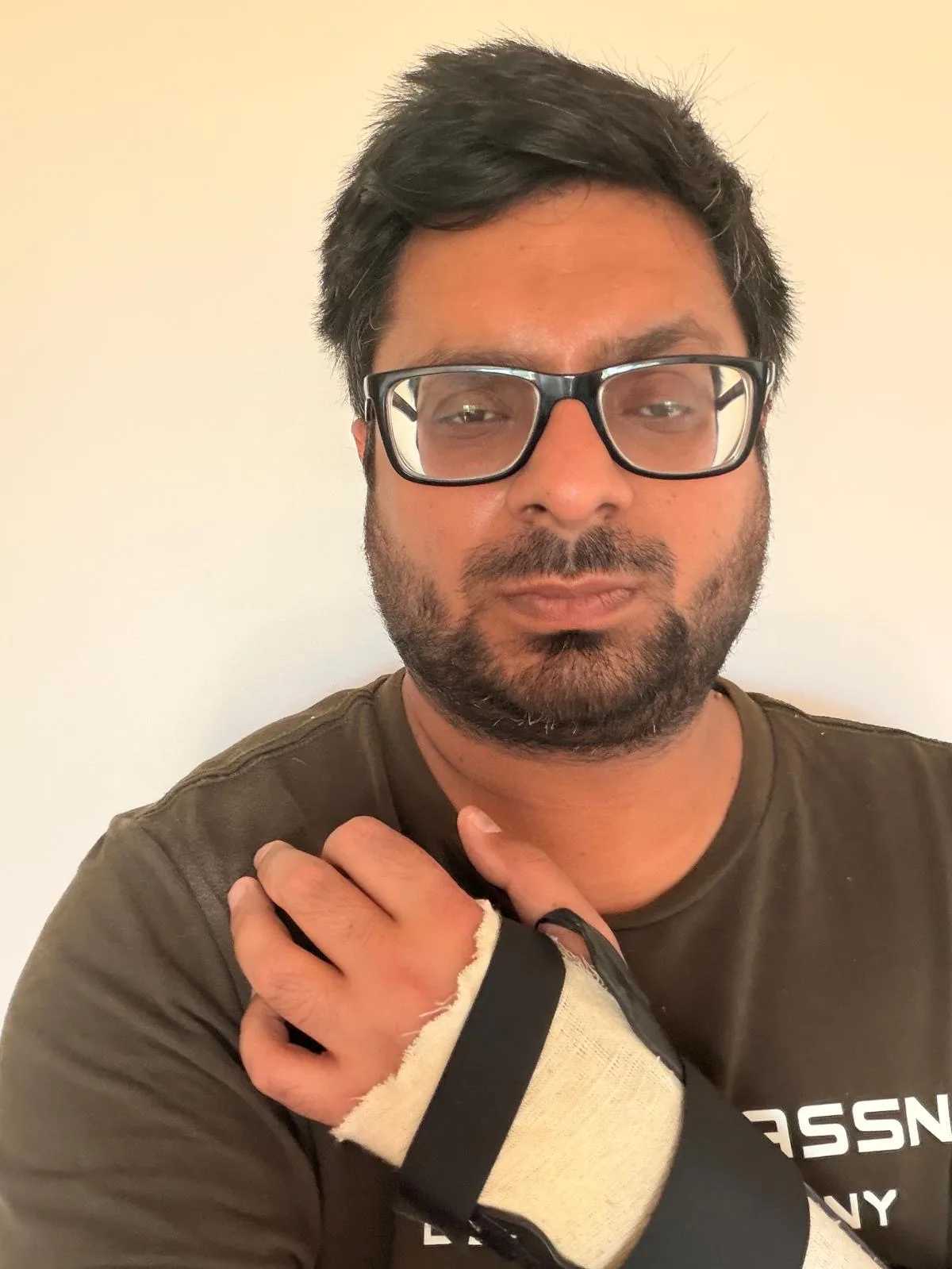 Man wearing glasses, holding up arm in a slingshot and looking into camera.