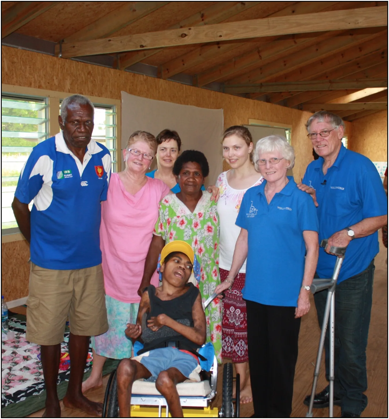 Two men and one woman wearing blue uniforms alongside several people who are smiling behind boy in a wheelchair. 