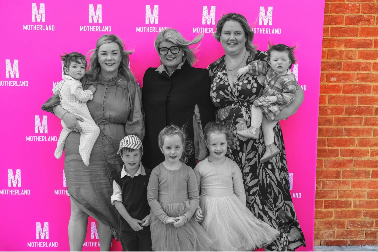 three women ,standing two holding children and threee children stood in fornt of a pink background reading Motherland
