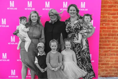 three women ,standing two holding children and threee children stood in fornt of a pink background reading Motherland