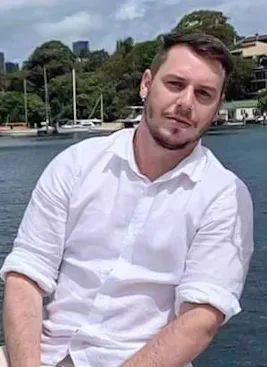 Man with short hair wearing long sleeve white button up shirt, sitting up and looking into camera.