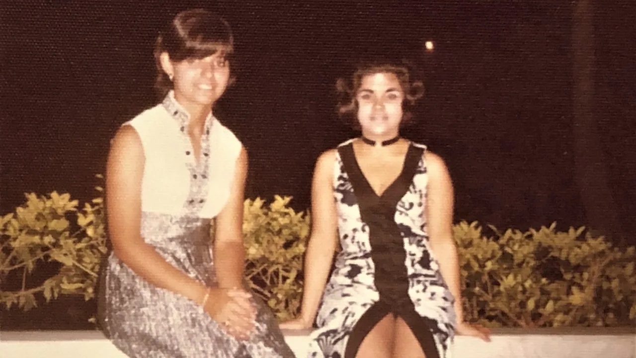 Two women wearing white and black dresses with shirt hair sitting on top of a pavement and smilling at camera.