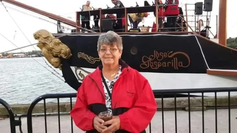 Elderly woman wearing rea jacket and glasses standing in front of a ship deck and smiling at camera.