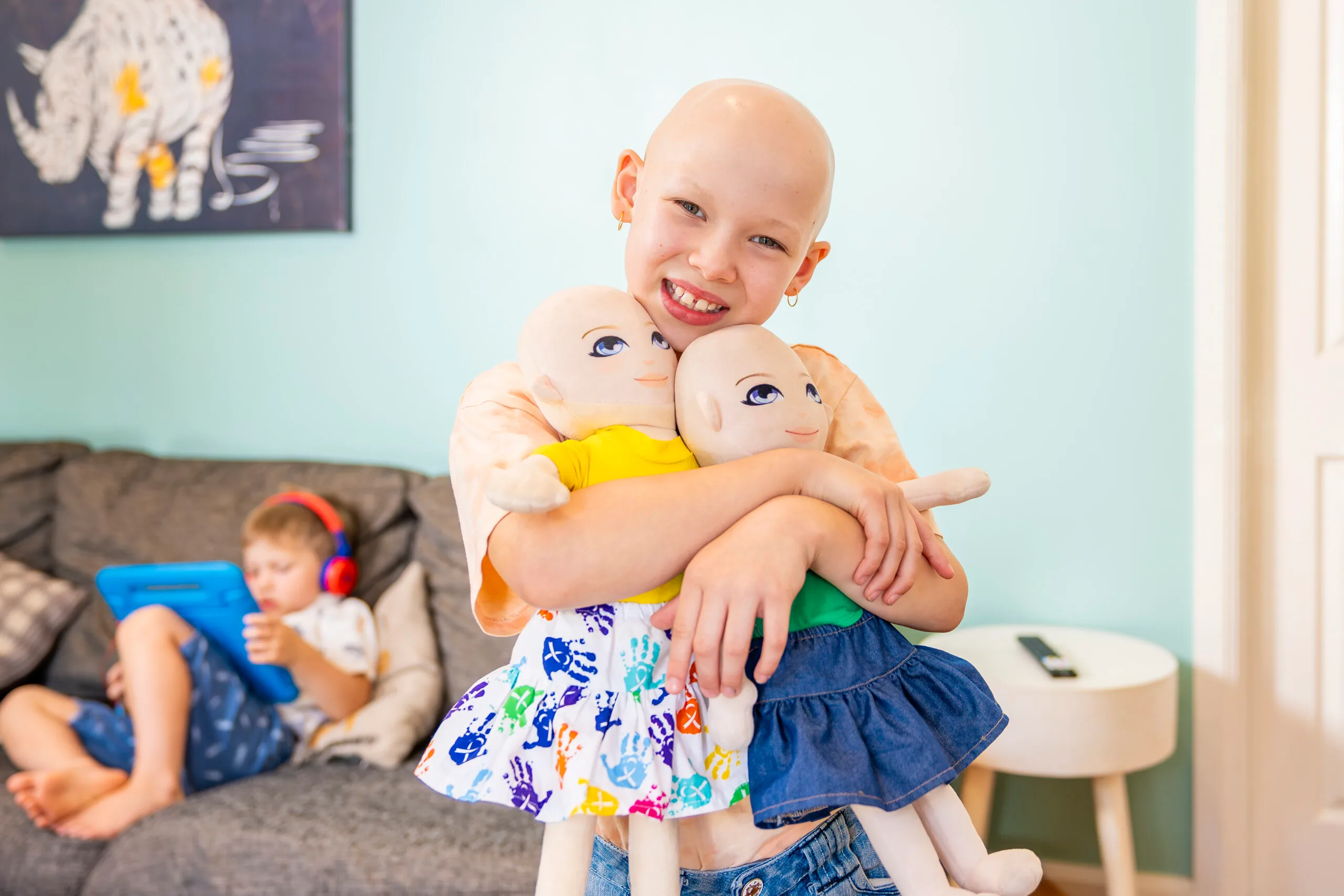 Little girl with no hair, wearing orange top and holding two dolls and smilling at camera.
