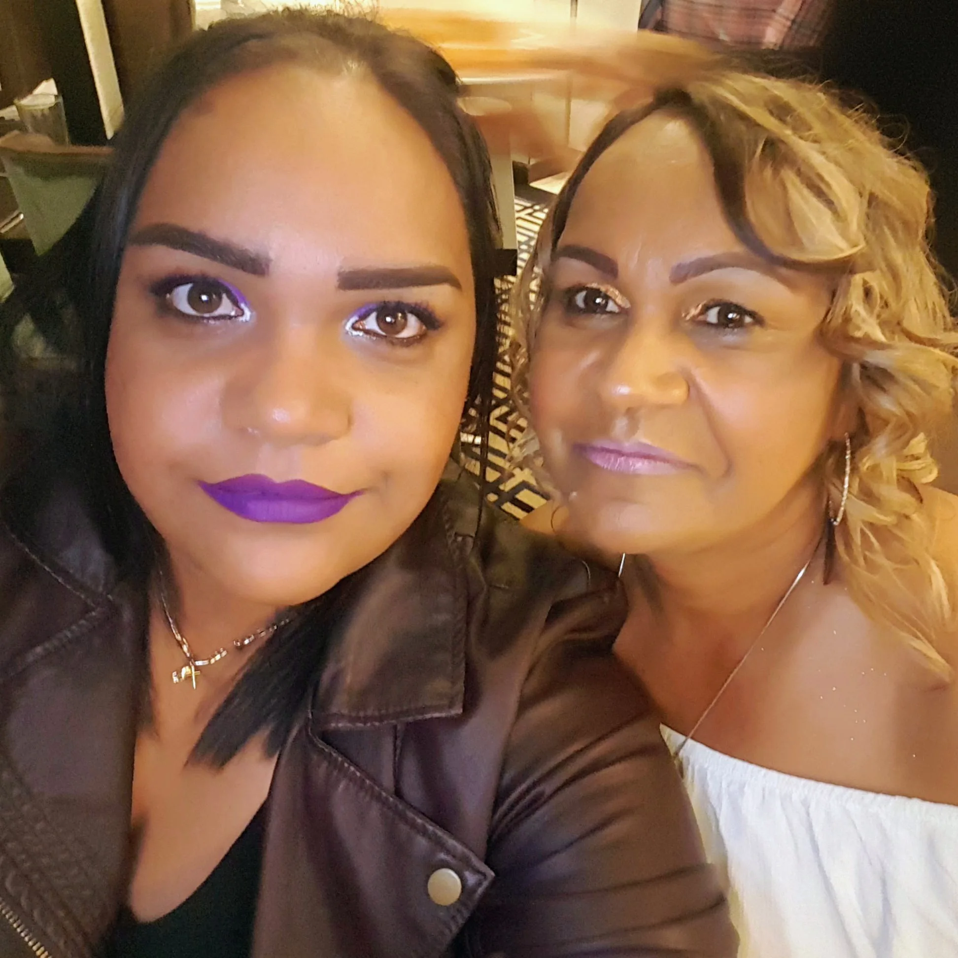 Woman with purple lipstick and glittery purple eyeshadow, dark hair next to woman with golden sparkly eyeshadow and blonde hair smilling at camera.