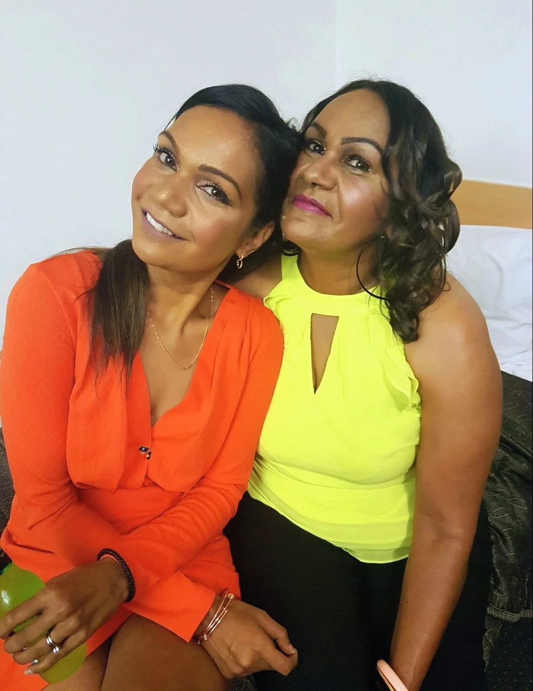 Woman wearing orange dress sitting next to woman wearing fleuro-yellow dress. both smilling at camera.
