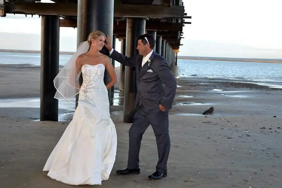A bride and groom stand under a pier on a beach