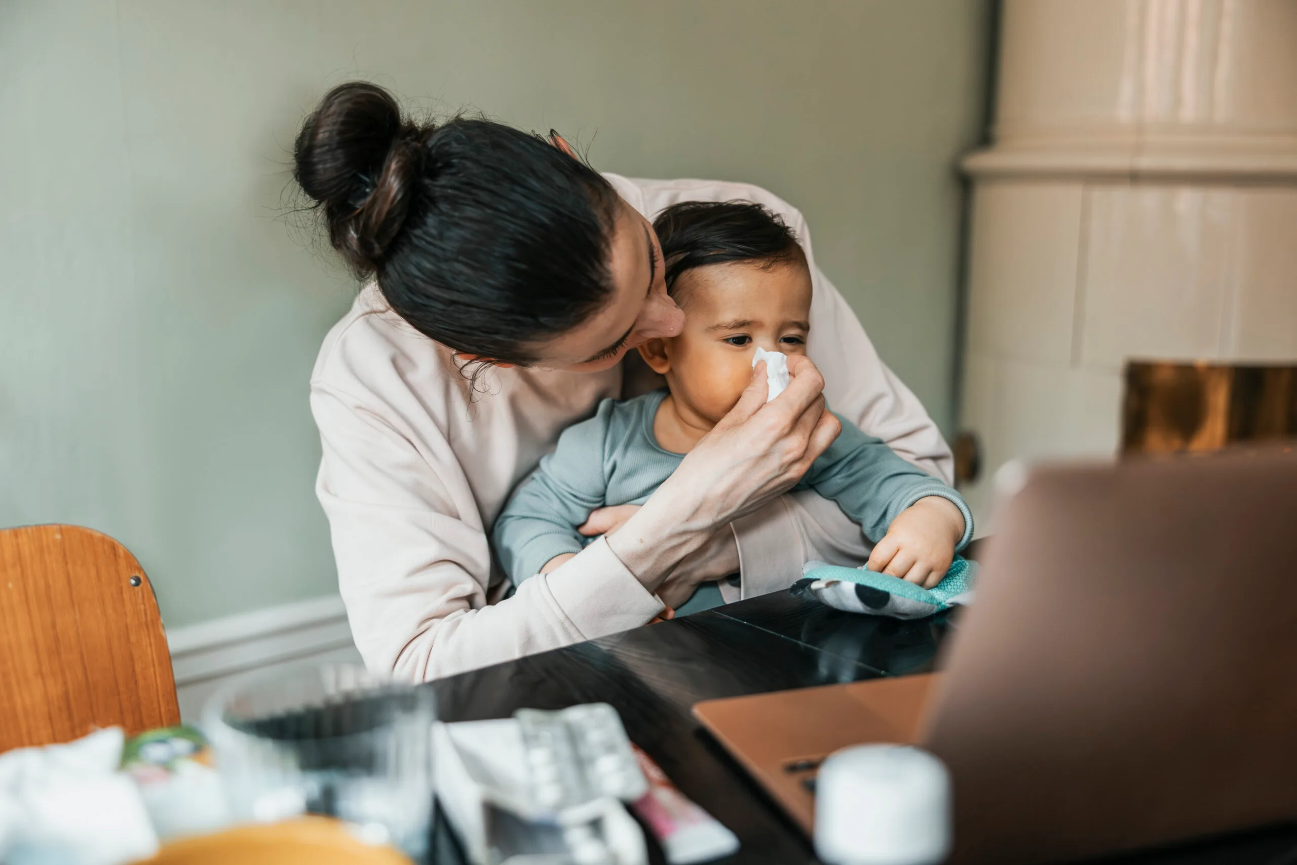Mum holding tissue to toddlers nose as they sit at a table at home