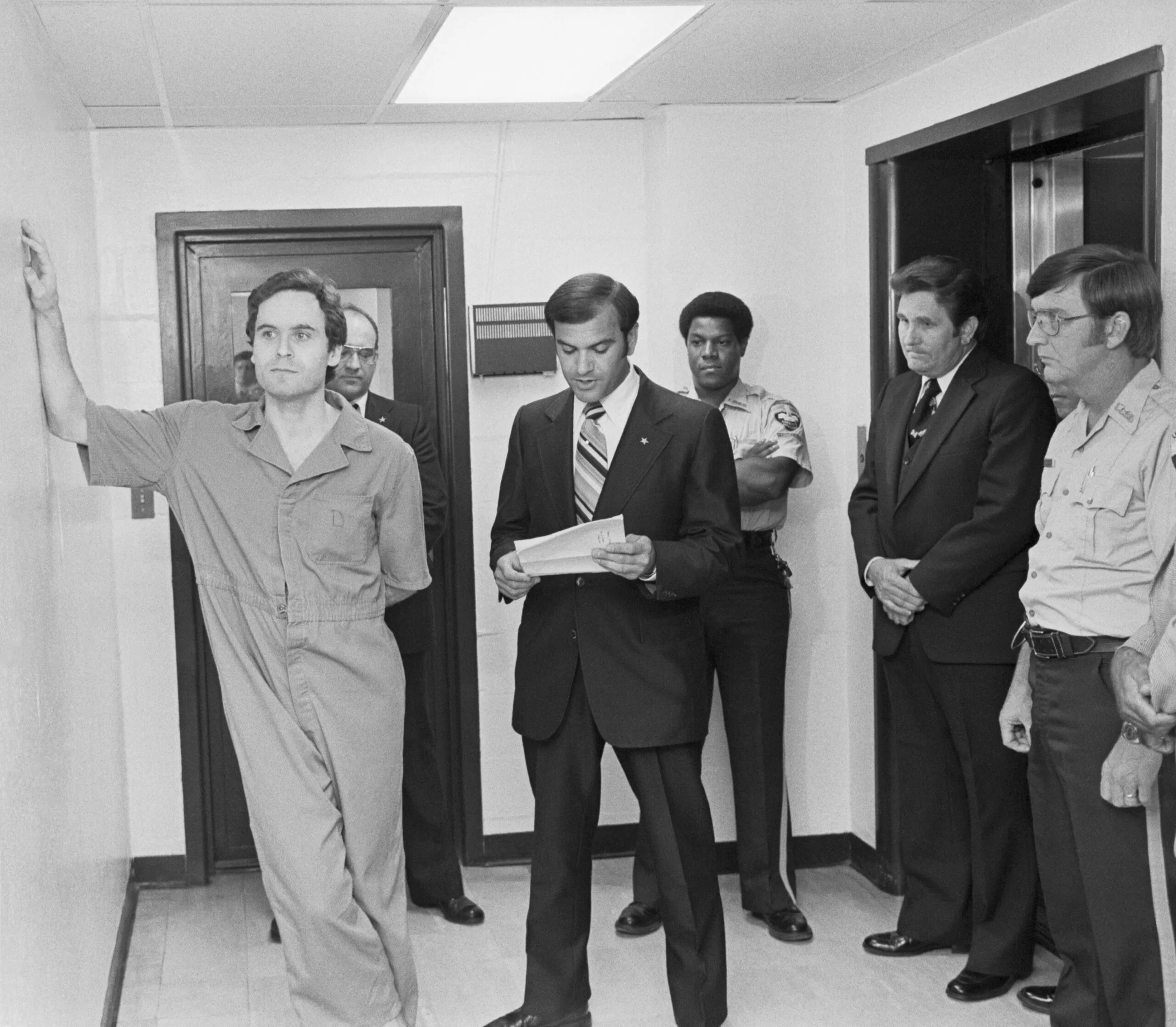Black and white photo of men dressed in suits, police uniform while speaking to another man in prisoner uniform.