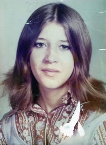 Picture of young girl wearing blue patterned shirt, with long brown hair, looking into camera.