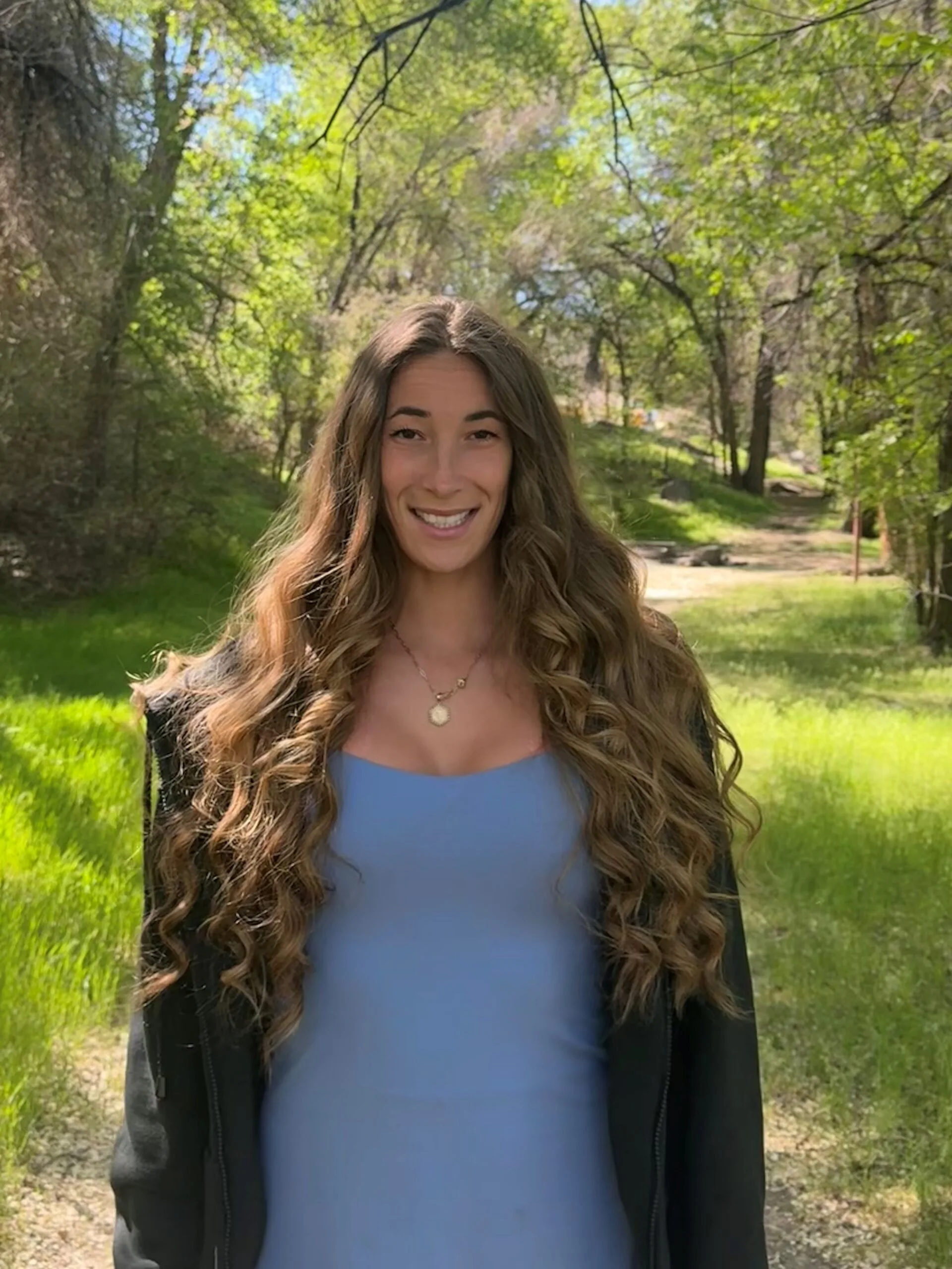 Woman with long brown curly hair, wearing blue top, standing in a park smilling at camera. 