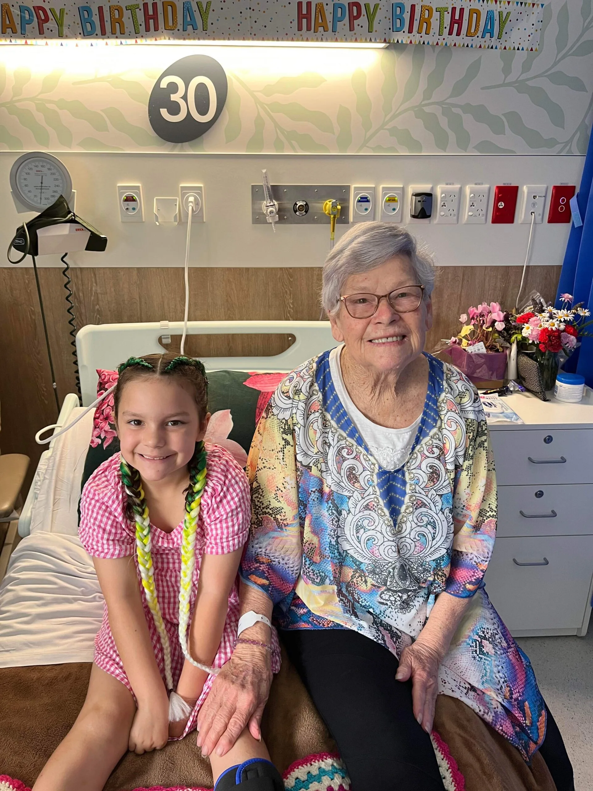 Elderly woman with grey hair, glasses and multicoloured top, sitting next to young girl wearing striped red dress. Both sitting on hospital bed and smilling at camera.