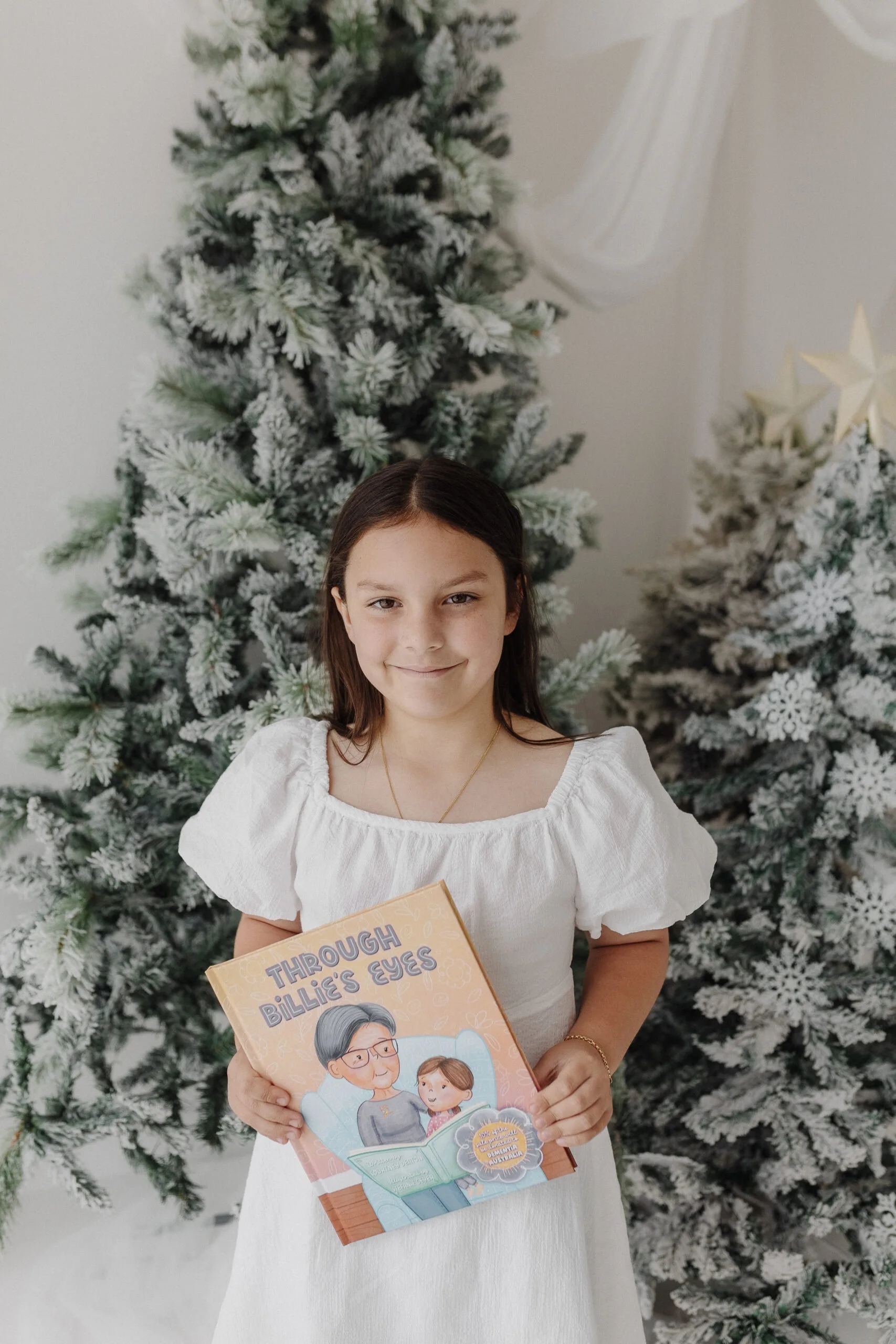 Young girl with long brown hair, wearing white dress, holding book and smilling at camera.