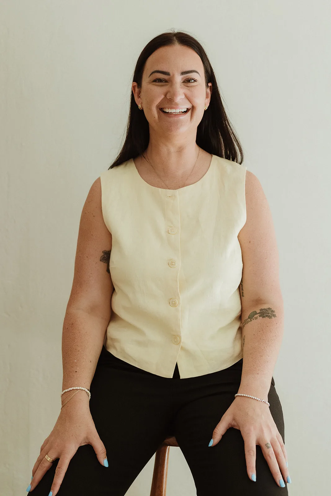 Woman with long brown hair, sitting on top of stool smilling at camera.