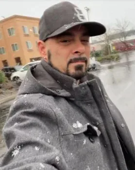 Man wearing grey jacket and black cap, smilling at camera.