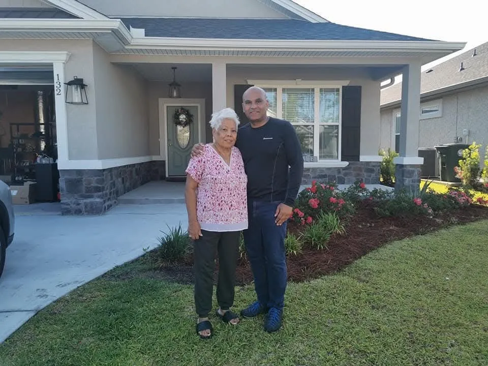 Woman wearing floral top and black pants standing next to man wearing black top and blue jeans, in front patio of house.