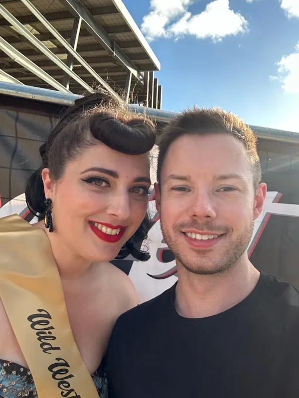 Woman with dark hair, wearing vintage dress with pageant sash, smilling next to man with short brown hair.