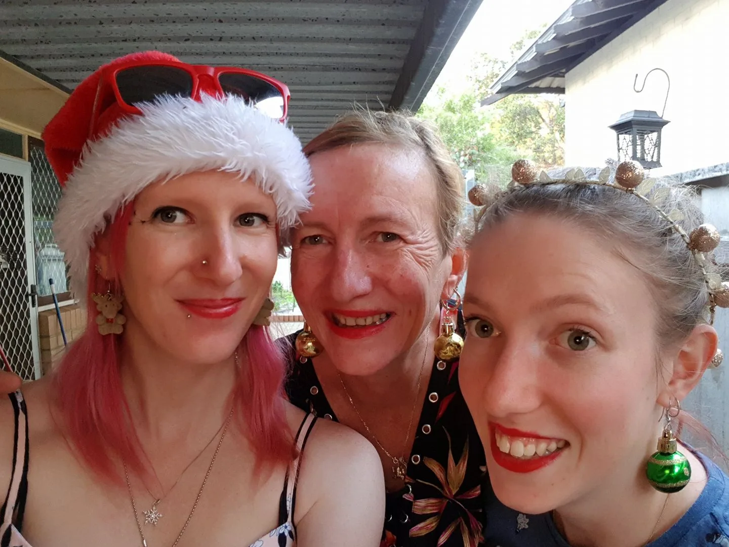Woman with pink hair wearing Christmas hat smiling next to another woman with blonde hair who is next to another woman wearing Christmas jewellery.