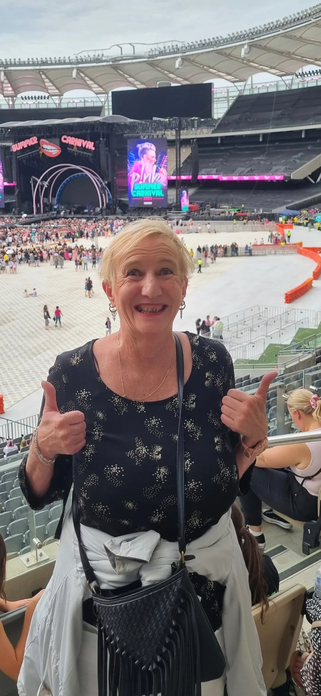 Woman with blonde hair, wearing black top, standing in a stadium and posing infront of camera with two thumbs up.