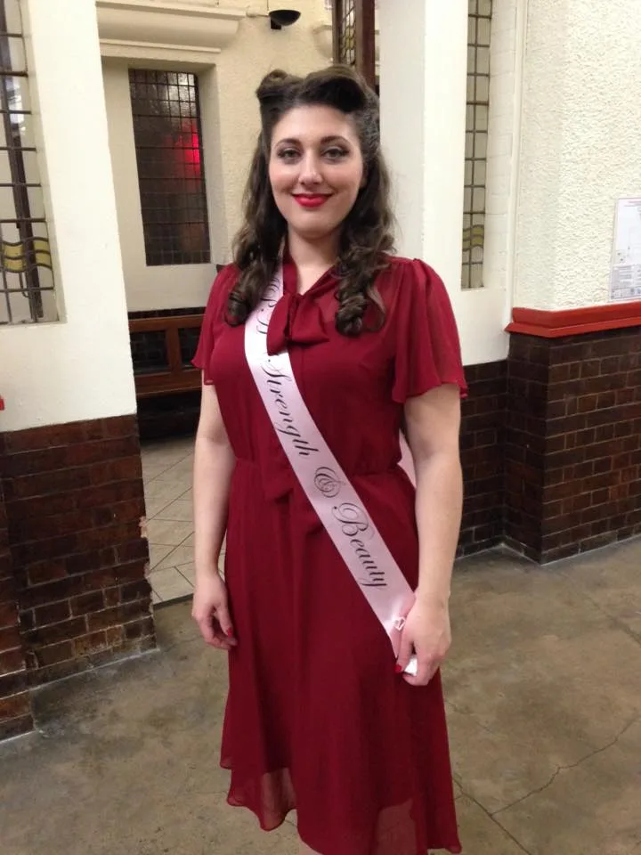 Woman with long brown hair, wearing long red dress with pageant sash across and smilling at camera.