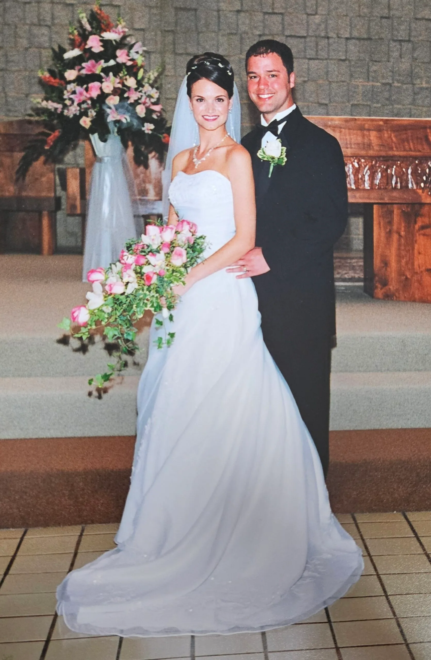 Woman in white wedding dress standing next to man in black suit at a church. Both smilling.