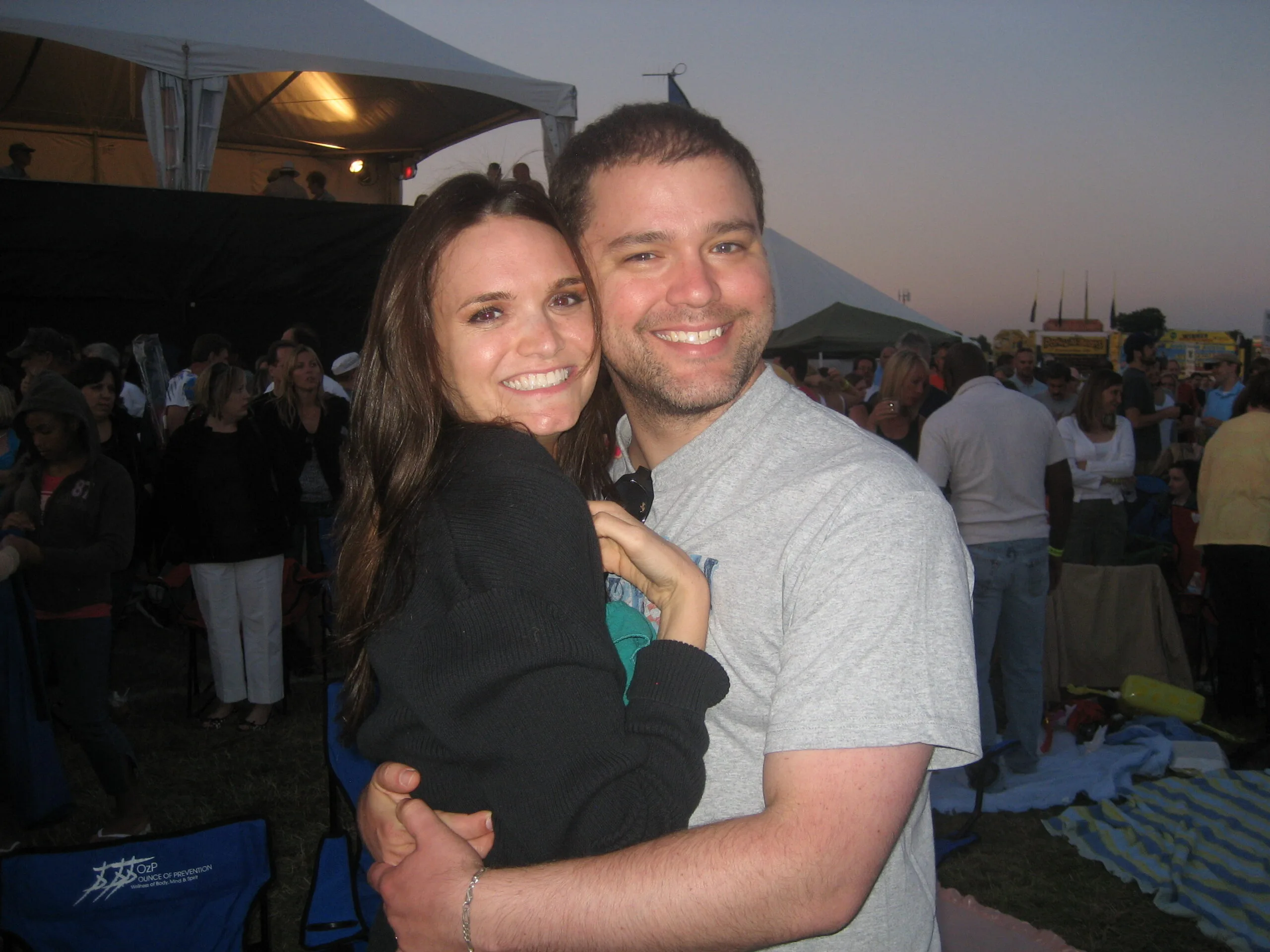 Woman wearing black jumper next to man in grey shirt at a festival, both smilling at camera. 