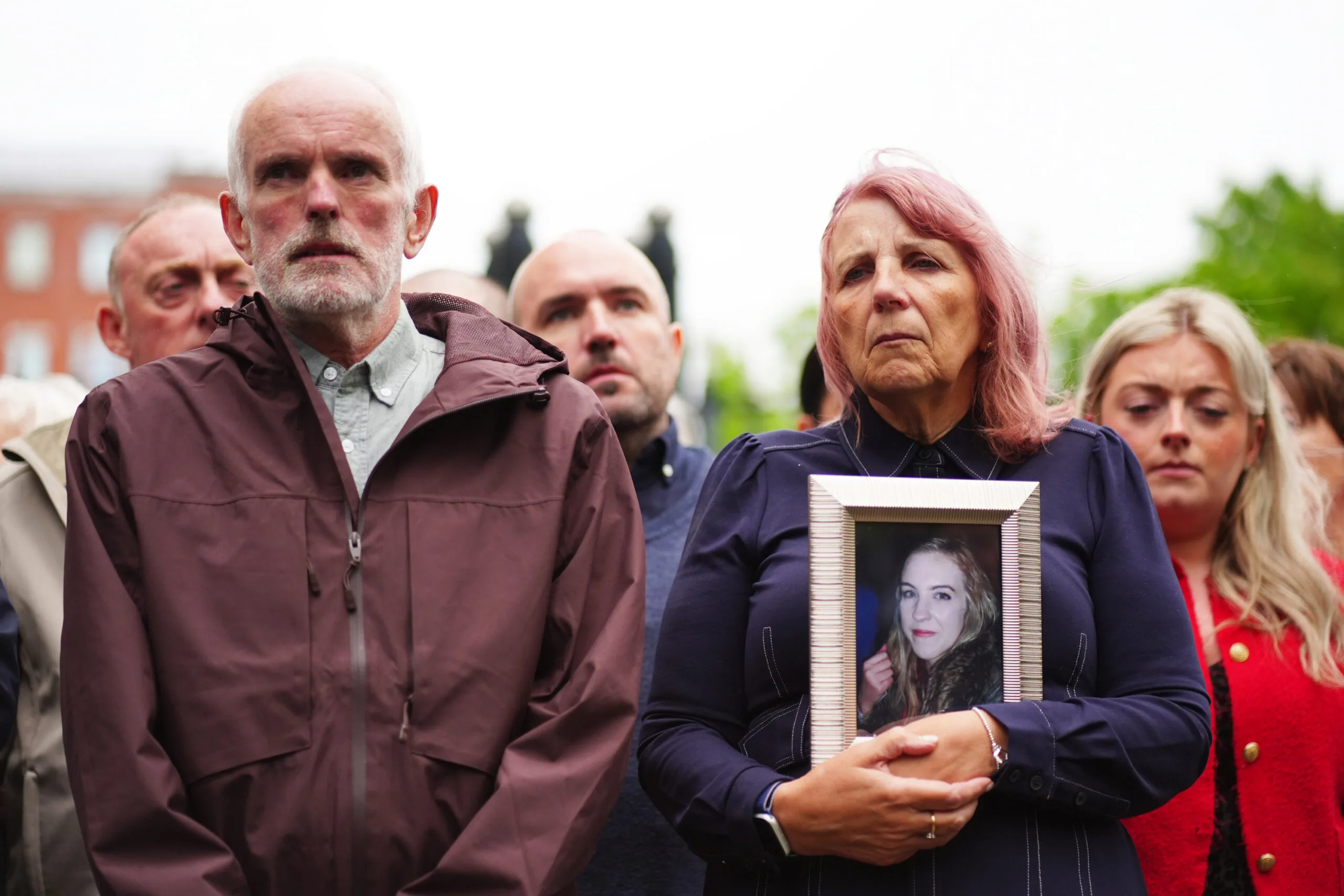 Man and woman holding up picture of woman with blonde hair. Both looking somber.