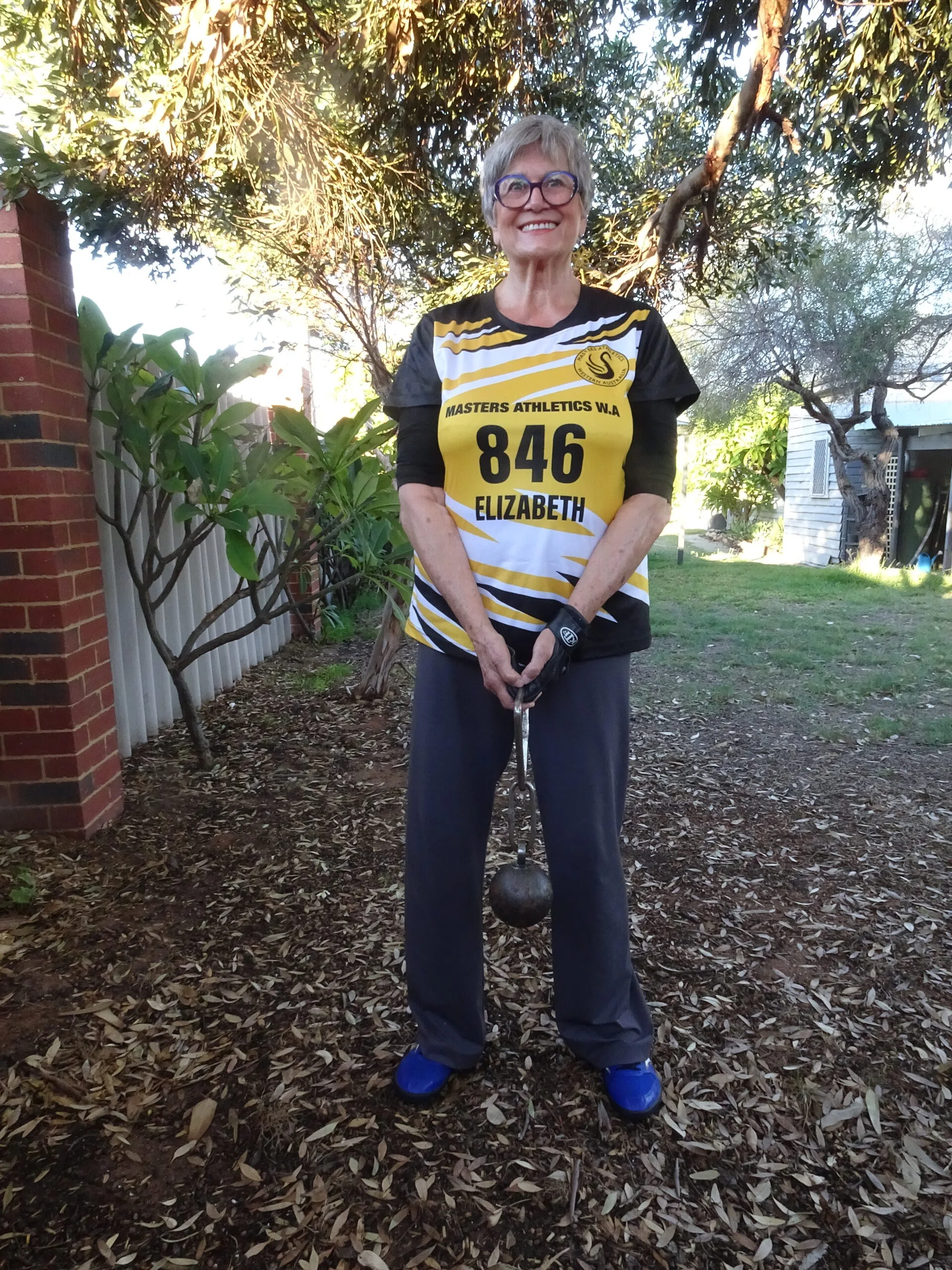 Woman wearing glasses, yellow sports top and pants, practising shot put in a backyard.