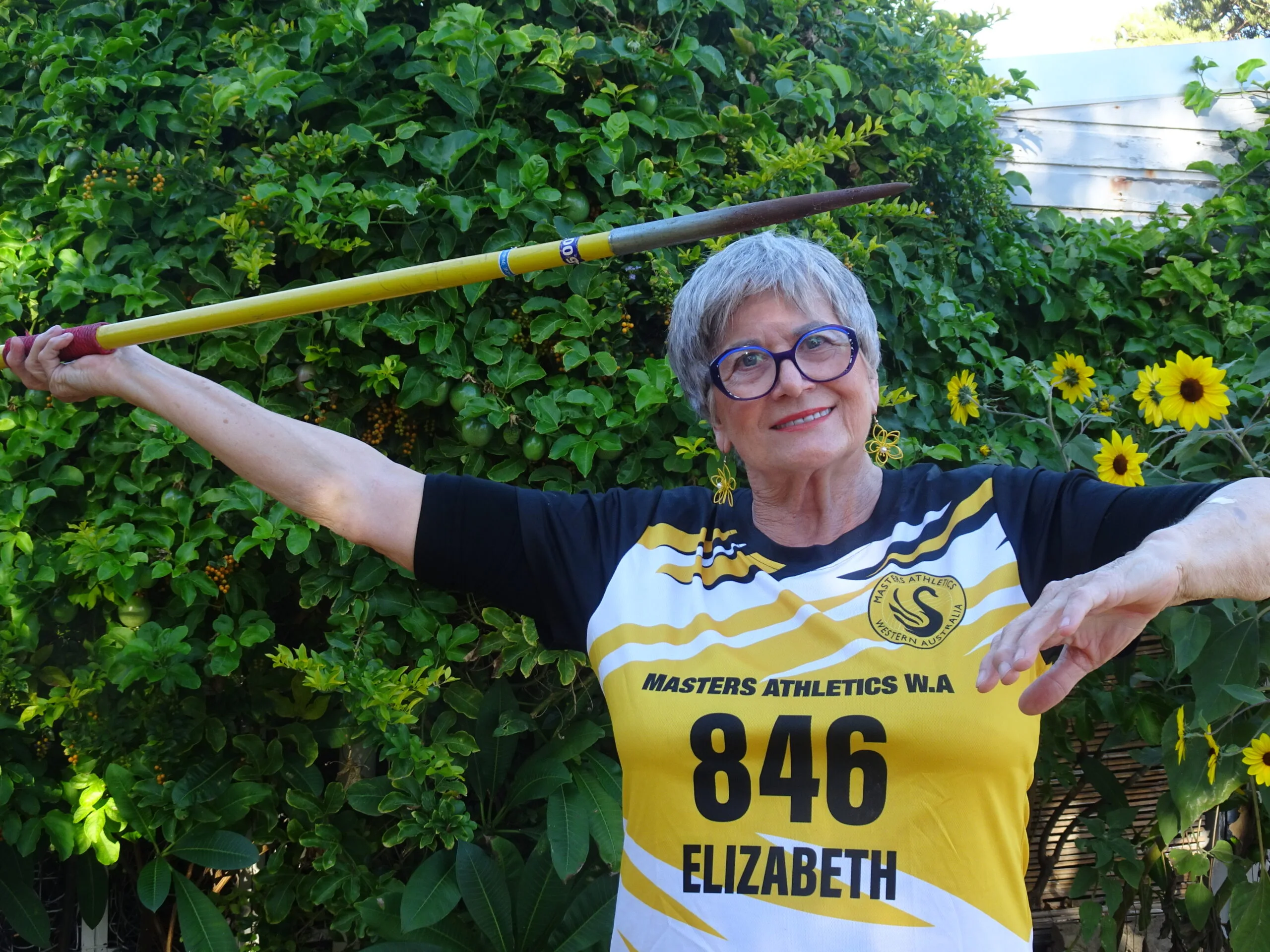 Elderly woman wearing glasses, a yellow athletic shirt and holding a javelin. 