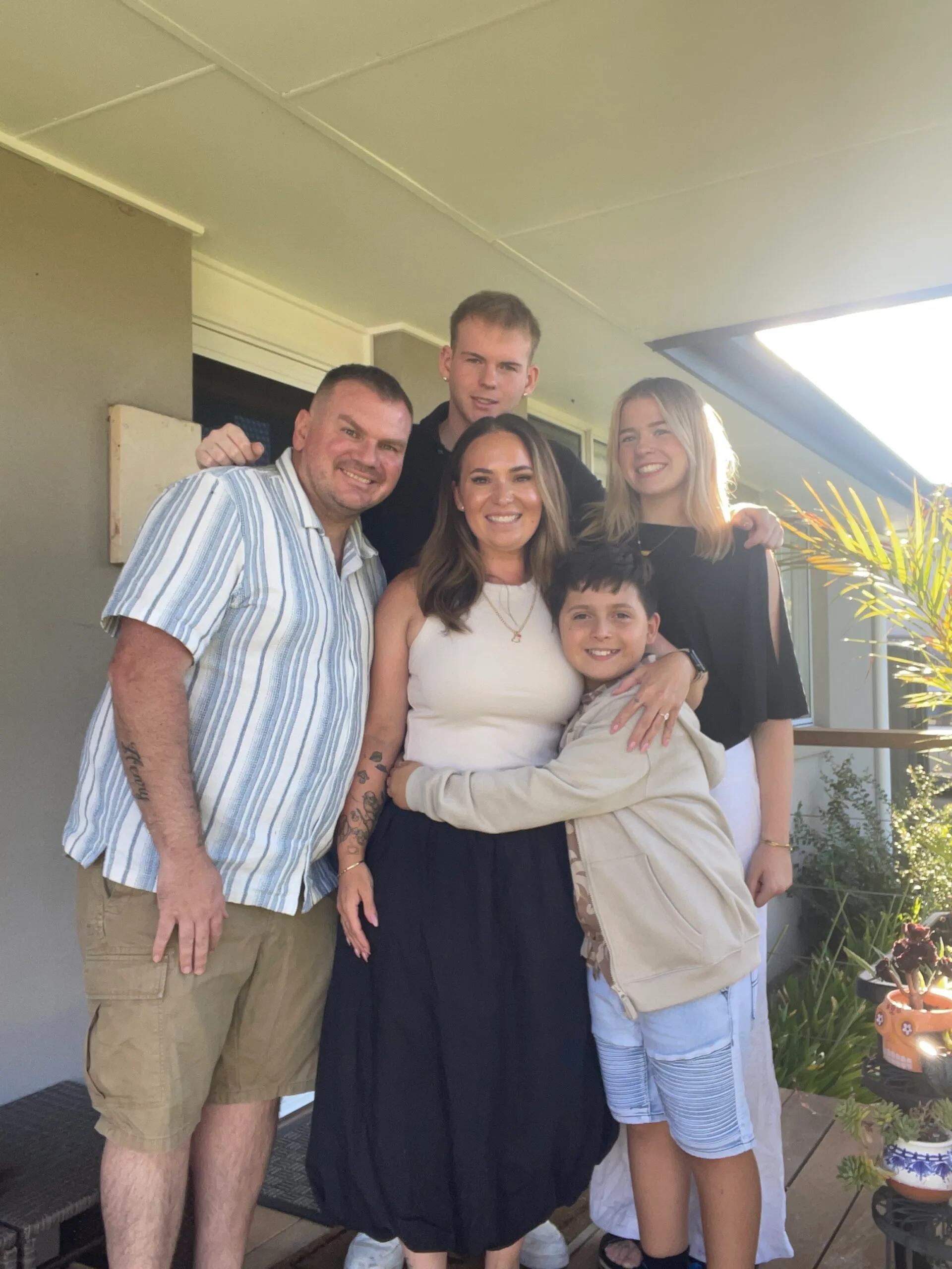Family photo of man wearing blue striped shirt, next to woman wearing beige top, next to young boy, girl and you man standing behind. All smilling at camera.