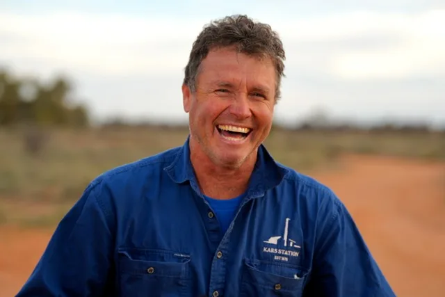 Man with grey hair, wearing a blue work shirt and smilling at camera in the outback.