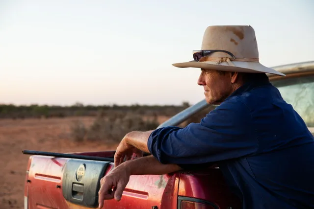 Man wearing blue shirt and cowboy hat leaning on red van and staring away from camera.