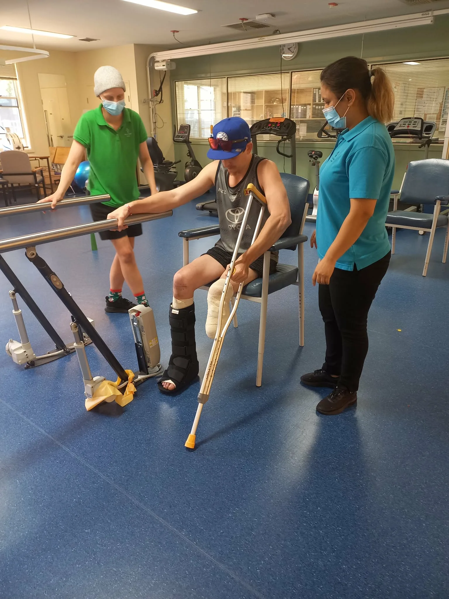 Man with amputated leg sitting on a blue chair holding on a railing, while woman and man stand next to him for support.