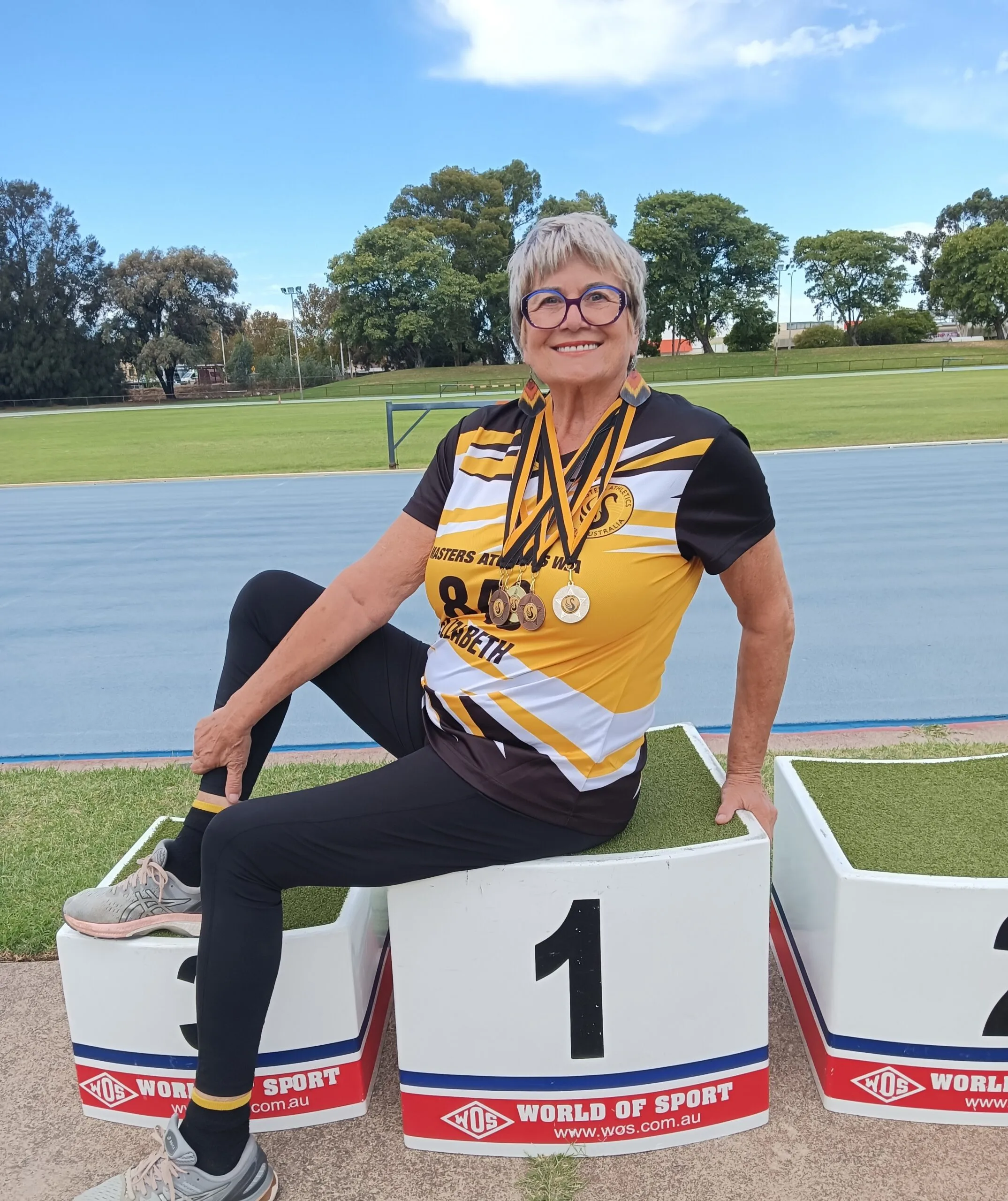 Woman wearing yellow sports top, with multiple medals sitting on top of position 1 block. 
