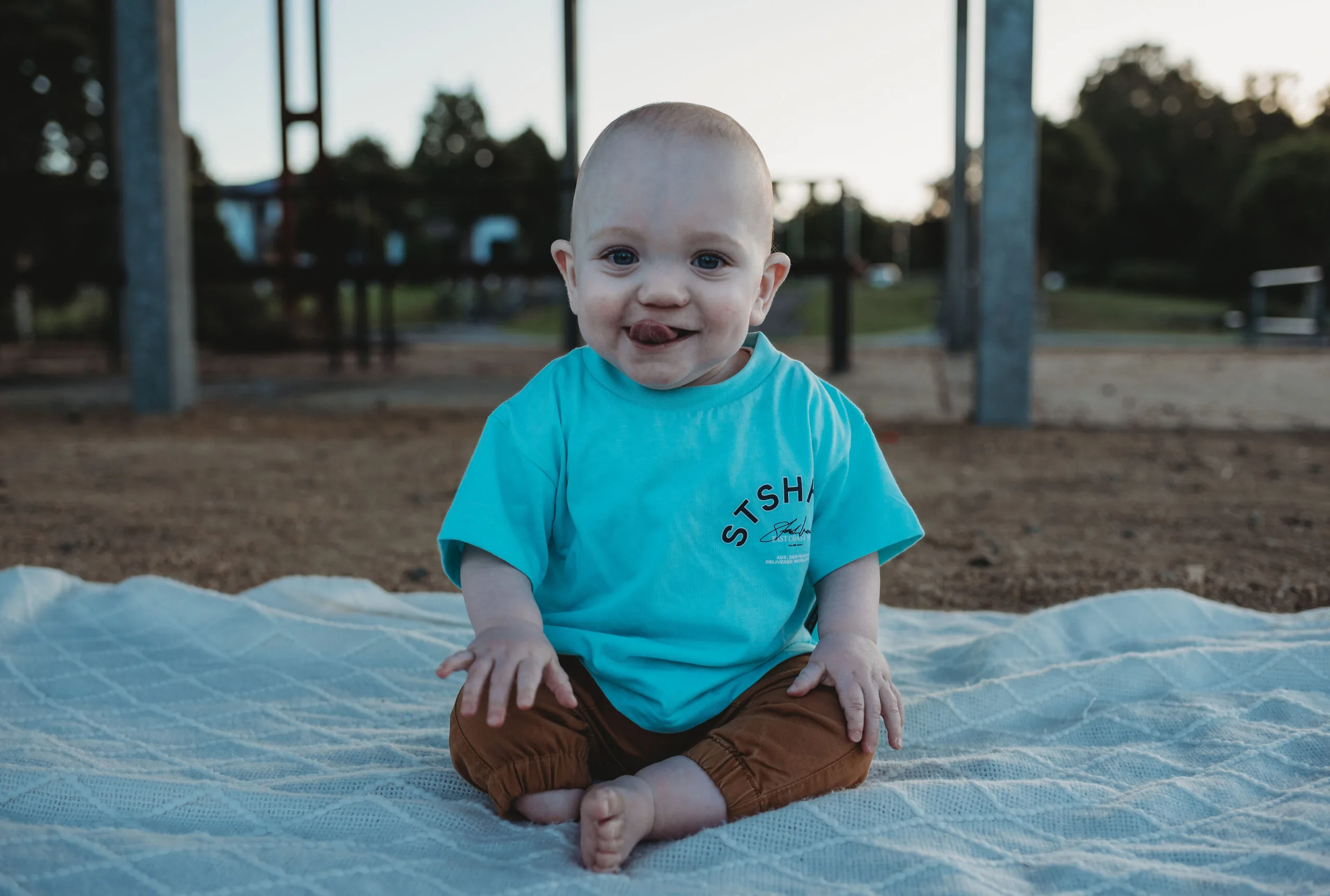 Baby wearing blue shirt and brown pants sitting on picnic blanket and smiling at camera.