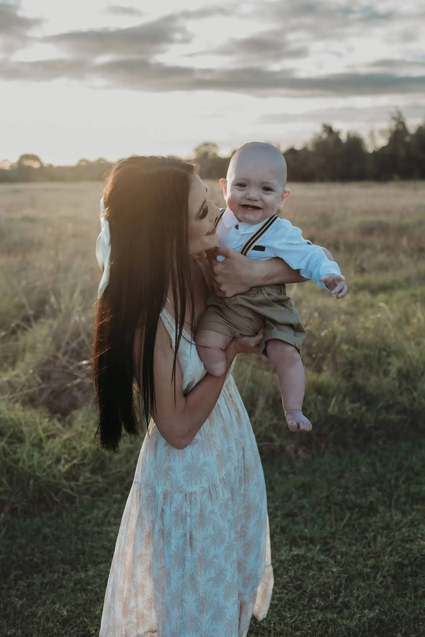 Woman in white dress holding up baby and standing in grass. Both smiling at camera.