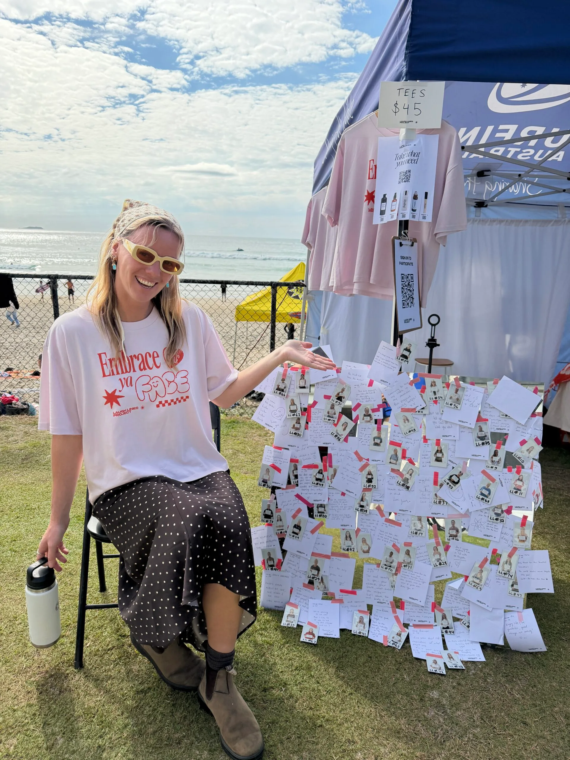 Woman wearing pick shirt, yellow sunglasses and bandanna and sitting next to stall pointing at photographs.