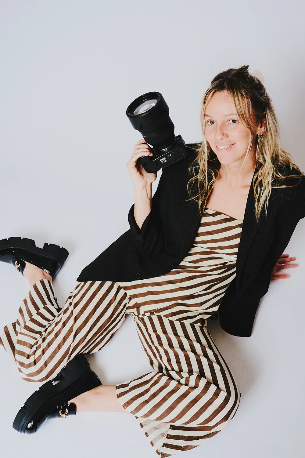 Woman wearing brown striped dress, holding up a camera and smiling.