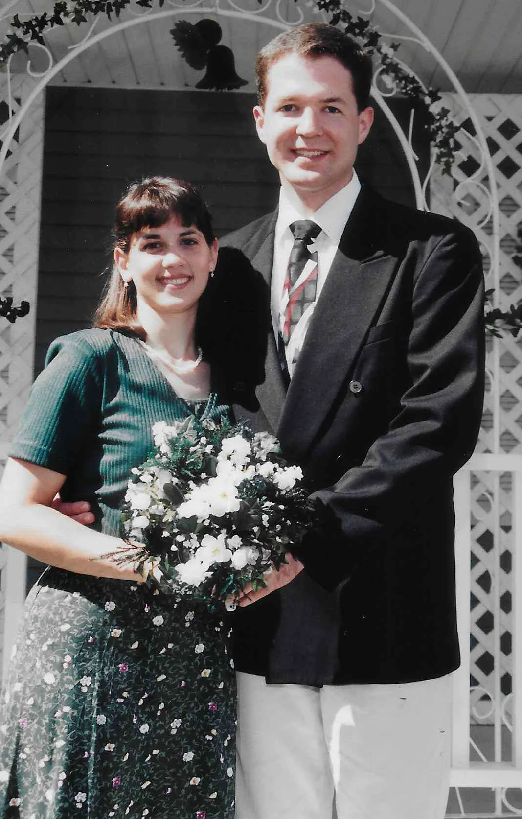 Woman wearing green dress holding flowers, standing next to man in a suit. Both smiling at camera.