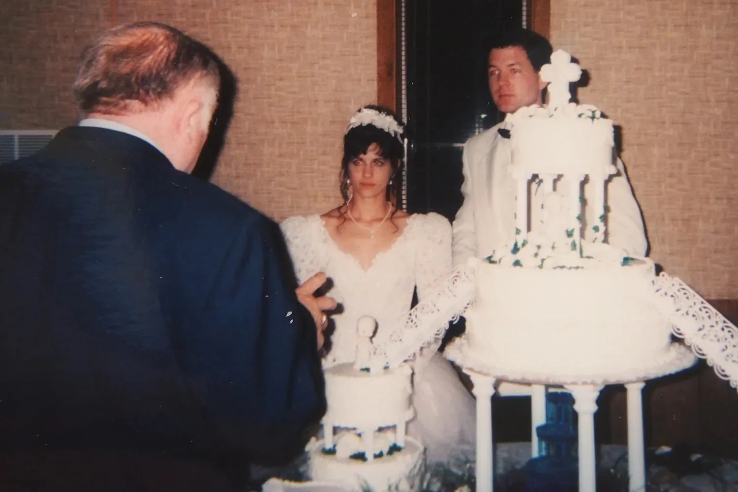Woman in wedding dress next to man in white wedding suit, both standing infront of wedding cake.