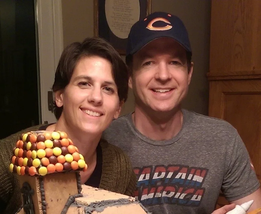 Man and woman smiling in front of homemade gingerbread house.