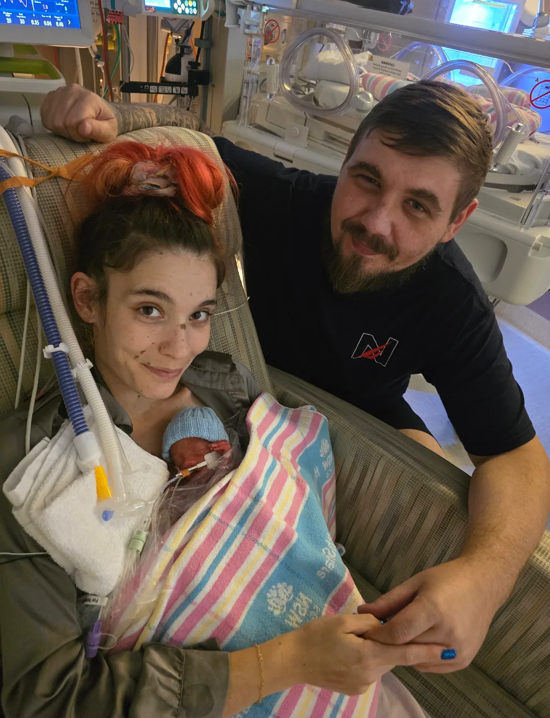 Woman in hospital chair, holding new born baby with man sitting next to her. Both smiling at camera.