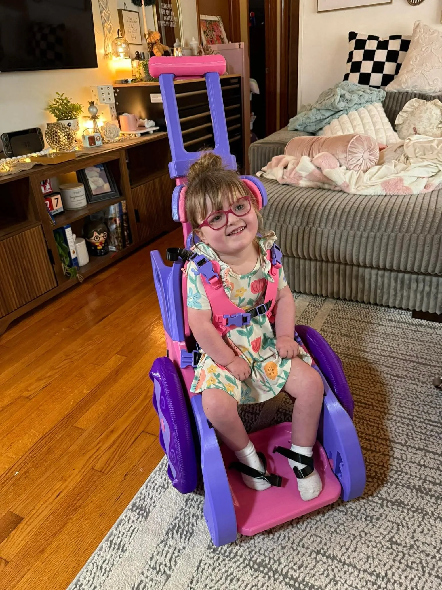 Little girl wearing red glasses and flowery dress, sitting in a purple and pink wheelchair.