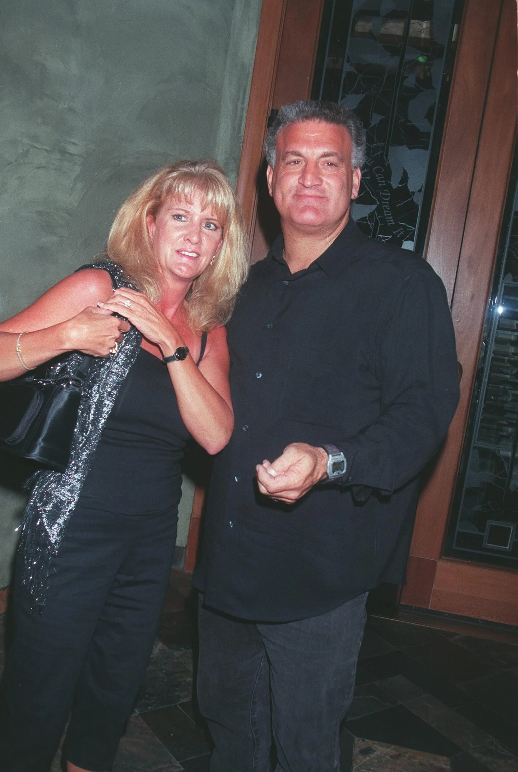 Woman with blonde hair, wearing black dress and black bag, standing next to man wearing black shirt and jeans.