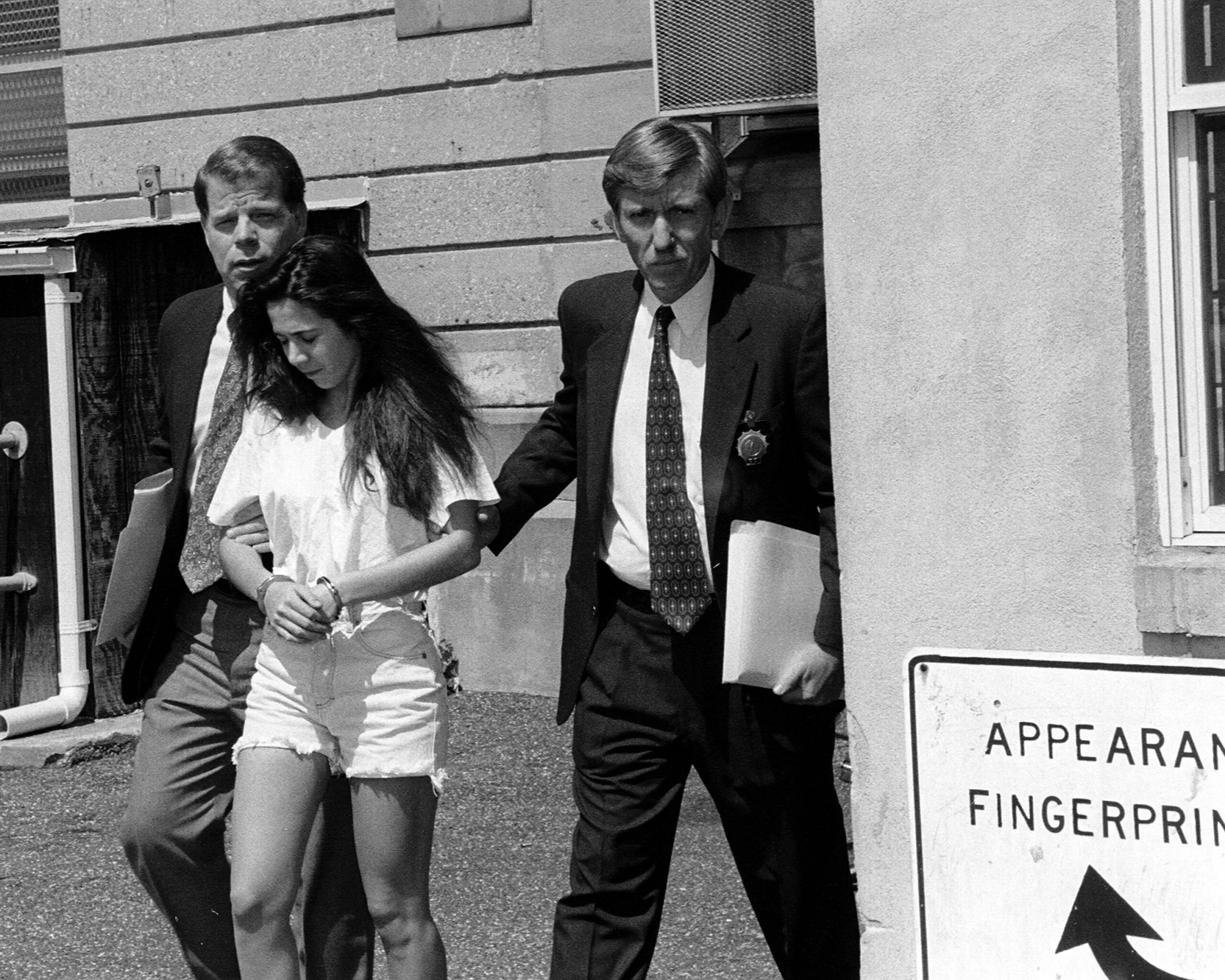 Black and white photo of teen girl in handcuffs being escorted by two men in suits.