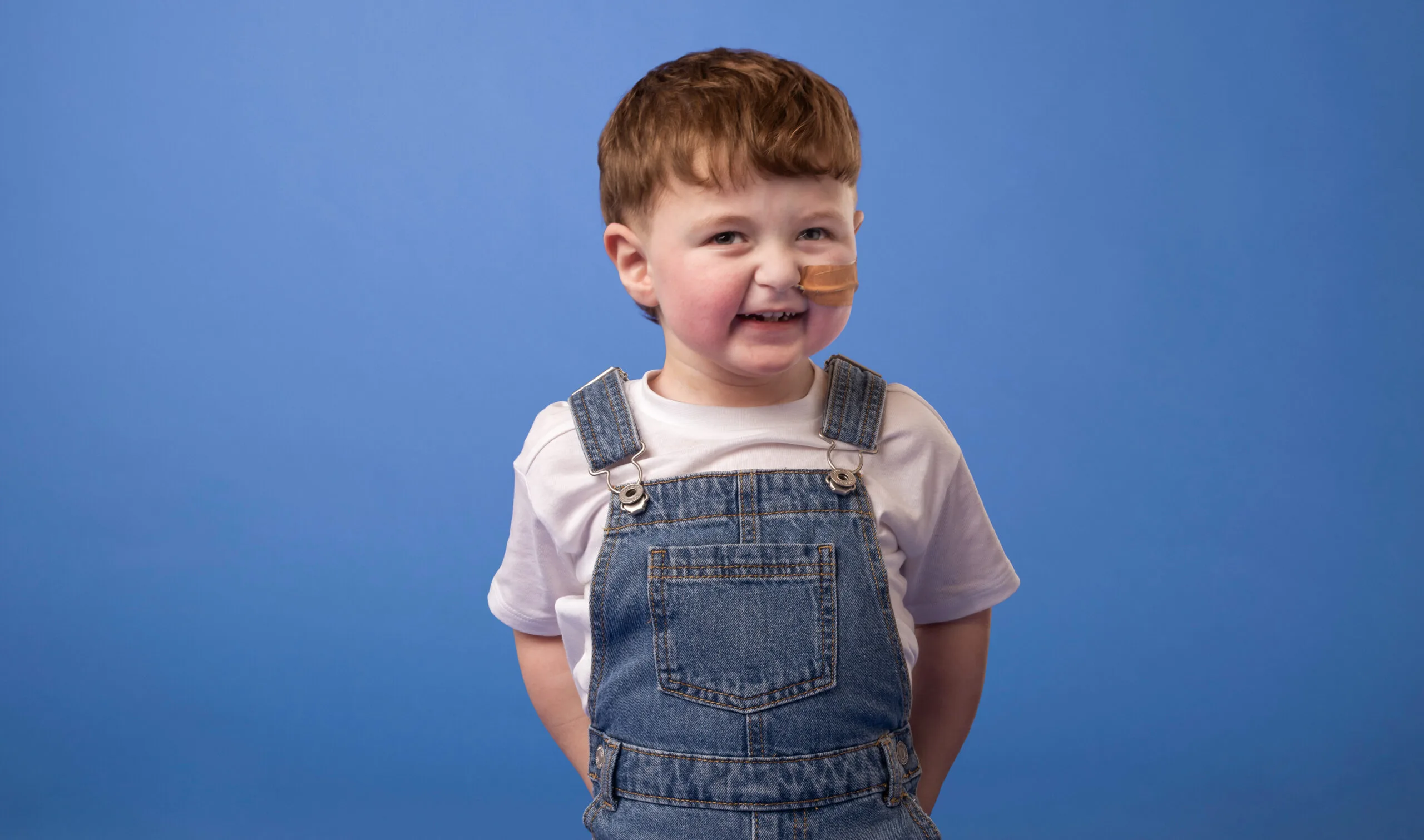 Toddler with feeding tube, wearing feeding tube and denim overalls, smiling at camera.
